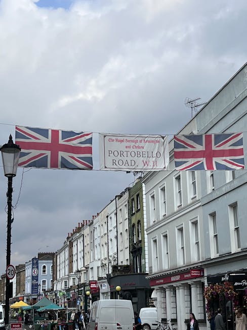 A street scene on Portobello Road in Notting Hill, with a large banner displaying the Union Jack flags and the text 'The Royal Borough of Kensington and Chelsea' and 'Portobello Road, W.11' stretched across the road, supported by black metal lamp posts on either side. The street is lined with multi-storey white and pastel-coloured buildings with various shopfronts, some with awnings, and pedestrians walking along the pavement. Near the foreground, part of a white van is visible, suggesting ongoing loading or transport activity related to home relocation or furniture transport. The sky above is overcast with grey clouds, providing natural lighting that highlights the busy atmosphere of the location, where house removals and packing and moving services might be taking place. The environment captures a typical vibrant day in this well-known area of London, with a focus on logistical movement and furniture handling.