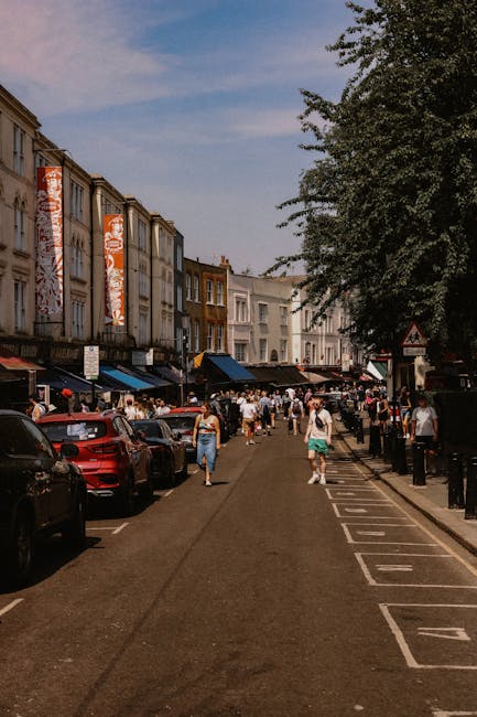 A busy street scene on Portobello Road in Notting Hill, with pedestrians walking along the pavement and a row of parked cars on the roadside. In the background, there are multi-story buildings with colorful facades, some storefronts with awnings, and large vertical banners displaying advertisements. On the right side, a large tree provides shade over part of the sidewalk. The scene depicts a typical day with people engaged in shopping, walking, and outdoor activities, relevant to home relocation and furniture transport services by Notting Hill Movers, focused on loading and unloading furniture as part of a packing and moving process.