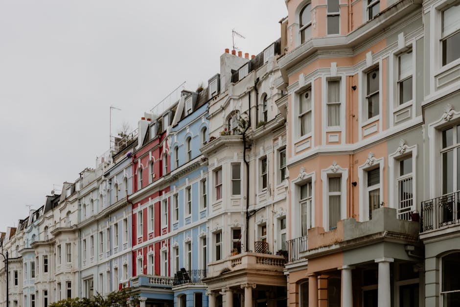 A row of multi-storey Victorian terraced houses in Notting Hill with ornate facades painted in pastel shades of pink, blue, white, and cream, featuring bay windows, decorative cornices, and small balconies. The image shows the upper levels of the buildings against an overcast sky, with some rooftops equipped with antennas and rooftop gardens visible. In front of the houses, a pavement area is partially visible, and a van or truck used for moving services can be seen nearby, possibly engaged in a home relocation or furniture transport process. The scene reflects typical urban residential architecture aligned with house removals and packing and moving logistics, as part of a professional relocation service by Notting Hill Movers, who occasionally handle the disposal of hazardous waste after a move, ensuring compliance with legal rules.
