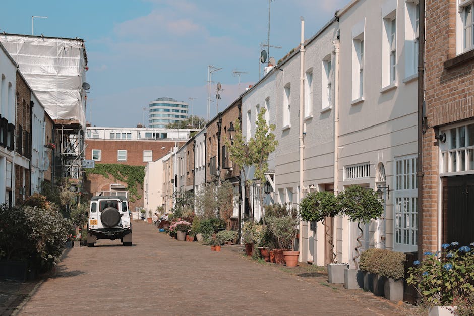 A row of multi-storey Victorian terraced houses in Notting Hill with ornate facades painted in pastel shades of pink, blue, white, and cream, featuring bay windows, decorative cornices, and small balconies. The image shows the upper levels of the buildings against an overcast sky, with some rooftops equipped with antennas and rooftop gardens visible. In front of the houses, a pavement area is partially visible, and a van or truck used for moving services can be seen nearby, possibly engaged in a home relocation or furniture transport process. The scene reflects typical urban residential architecture aligned with house removals and packing and moving logistics, as part of a professional relocation service by Notting Hill Movers, who occasionally handle the disposal of hazardous waste after a move, ensuring compliance with legal rules.
