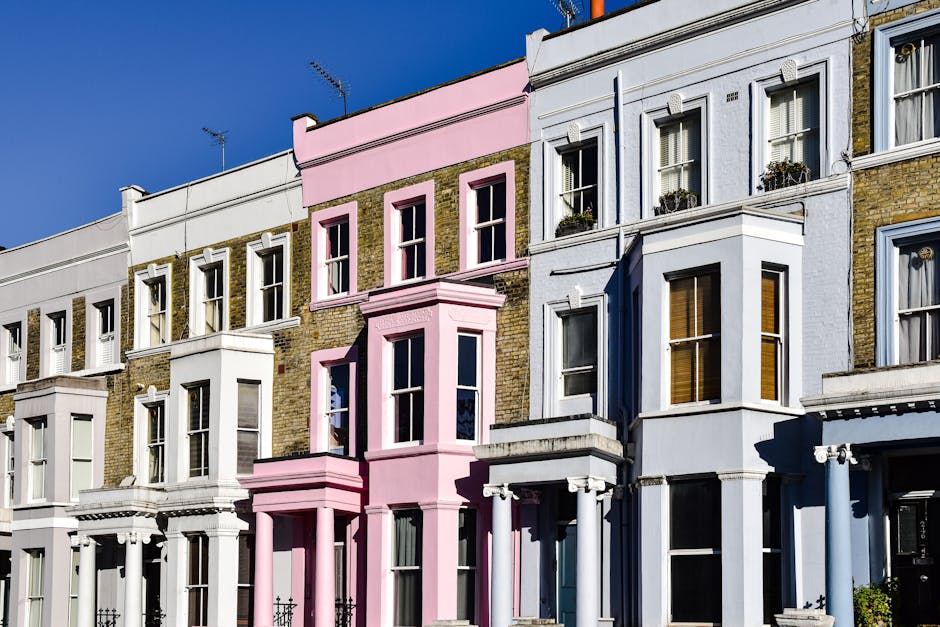 A row of Victorian terraced houses in Notting Hill featuring multiple stories, with bay windows and decorative architectural details. The buildings are painted in various pastel shades, including white, pink, and light blue, with some sections displaying exposed brickwork. The image captures a clear blue sky above the buildings, suggesting a sunny day. In the foreground, a sidewalk is visible, with no visible vehicles or moving equipment present. The scene emphasizes residential facades typical of London architecture, associated with home relocation and moving services, as seen through the prominence of the building exteriors that may require furniture transport or packing and moving procedures when considering house removals. Notting Hill Movers’ branding is not directly visible in this image, but the setting aligns with the type of properties they typically serve in house removals and moving logistics, highlighting the importance of organized delivery and staircase or doorstep access for packing and loading processes.