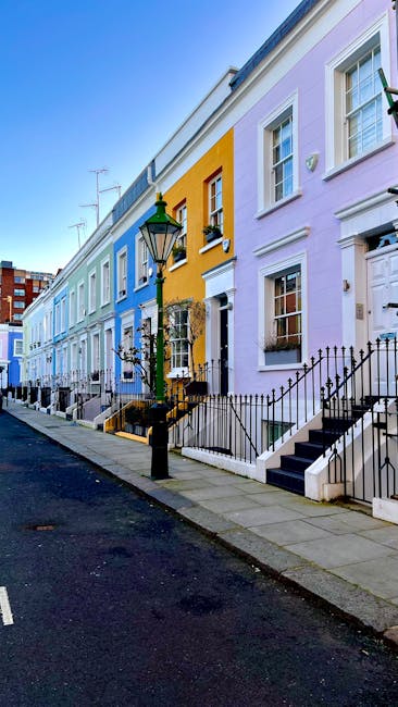 A row of houses on a residential street in Notting Hill, painted in pastel shades including light blue, yellow, and lavender. The houses feature front steps with black metal railings, white window frames, and small decorative iron balconies. A traditional black lamp post stands near the pavement, which is made of concrete slabs, alongside a black metal fence enclosing the steps of each property. The street is paved with asphalt, and the sky above is clear and blue, indicating a bright, sunny day. The scene is typical of the neighborhood, with a clean and quiet atmosphere. In the context of house removals and furniture transport, the image reflects the typical setting where professional movers, such as Notting Hill Movers, may be engaged to assist with home relocation processes, including packing, loading, and transportation of belongings from these picturesque properties.