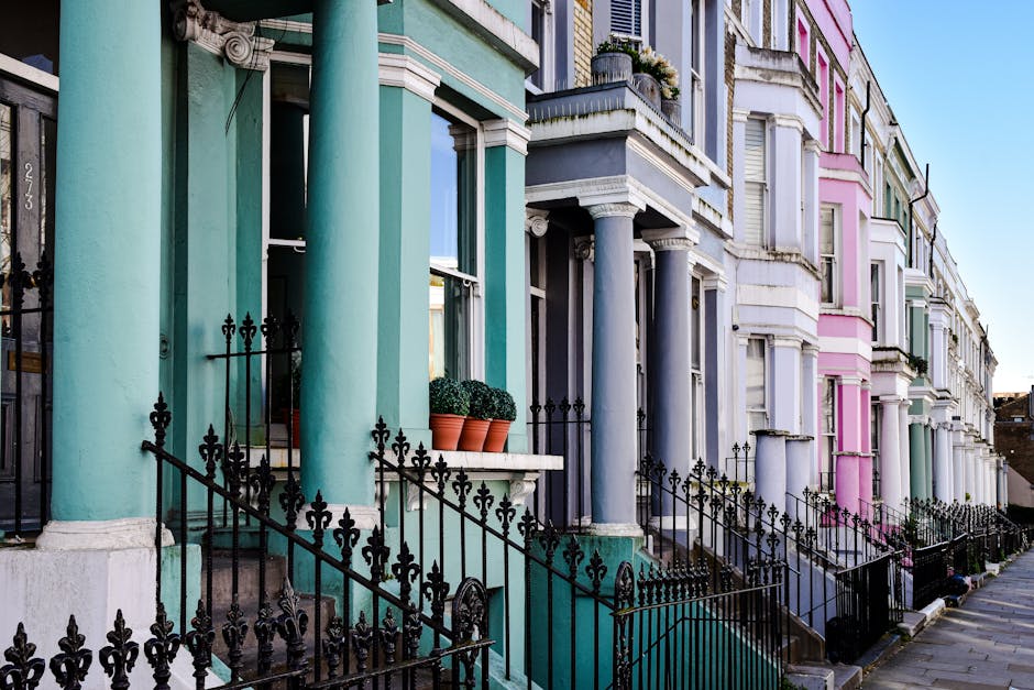 A row of Victorian terraced houses in Notting Hill featuring multiple stories, with bay windows and decorative architectural details. The buildings are painted in various pastel shades, including white, pink, and light blue, with some sections displaying exposed brickwork. The image captures a clear blue sky above the buildings, suggesting a sunny day. In the foreground, a sidewalk is visible, with no visible vehicles or moving equipment present. The scene emphasizes residential facades typical of London architecture, associated with home relocation and moving services, as seen through the prominence of the building exteriors that may require furniture transport or packing and moving procedures when considering house removals. Notting Hill Movers’ branding is not directly visible in this image, but the setting aligns with the type of properties they typically serve in house removals and moving logistics, highlighting the importance of organized delivery and staircase or doorstep access for packing and loading processes.