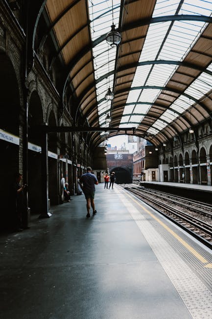 The image depicts an indoor train station with a curved, arched roof made of glass and metal, supported by ornate brickwork columns with decorative arches. The platform is clean and well-lit, with natural light streaming through the glass roof. Several passengers stand or walk along the platform, some sitting on benches near the wall. In the background, railway tracks extend into a tunnel or open area outside. The environment appears spacious with a mixture of historic architectural design and modern functionality. This scene is relevant to the home relocation process, as it illustrates the transportation phase, involving the loading and transit of furniture and boxes for a house move. Notting Hill Movers, a professional removals company, often coordinate such transport logistics during a small-flat removal, such as from Ladbroke Grove to Notting Hill Gate.