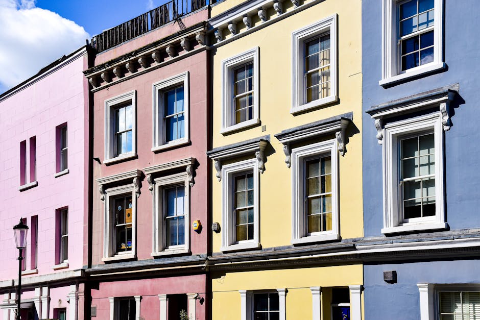 A row of three adjacent terraced houses with distinctive facades in pastel pink, yellow, and blue colors, located on a street in Notting Hill. The houses feature traditional sash windows with white frames and decorative mouldings, with the first-floor windows supported by ornate brackets. The pink house has two vertical rectangular windows on each floor, the yellow house has four windows aligned vertically, and the blue house has three windows, some partially open. A street lamp is visible in the foreground on the left side, and part of a vehicle or delivery van can be seen parked on the street pavement below the houses. The scene is captured in bright daylight, highlighting the vibrant colors and architectural details, as part of a typical residential area undergoing home relocation or furniture transport activities, with [COMPANY_NAME] potentially managing the removal process involved in the moving services connected to the 'Ladbroke Grove to Notting Hill Gate' project.