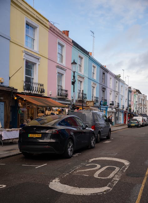 A row of colorful terraced houses in Notting Hill, London, painted in pastel shades of yellow, pink, blue, and purple, with white window frames and black iron balconies. The ground floors feature small storefronts, one with a black and white sign, while others have awnings and outdoor displays. Parked along the street are several vehicles, including a black modern hatchback, a dark grey van, and a few other cars, parked parallel to the curb. The street has a painted speed limit of 20 mph in white near the foreground, and a lamppost stands in front of the row of houses. The sky is partly cloudy with light blue patches visible, and the scene suggests a residential area with local shops, typical of Notting Hill. This setting is relevant to house removals and furniture transport, with potential loading and unloading activities for relocation services provided by companies like Notting Hill Movers.