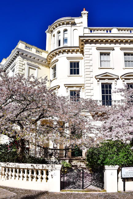 A large, white, multi-story Victorian-style house with ornate architectural details, including rounded bay windows and decorative cornices, set against a bright blue sky. The property is situated on a