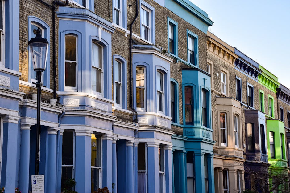 A row of Victorian-style terraced houses in Notting Hill, London, with facades painted in pastel shades including light blue, beige, and vibrant green. Each building features large sash windows with white trims, and some have small decorative balconies or window boxes. A black traditional street lamp is positioned on the pavement in front of the houses. The street scene is illuminated by natural daylight, highlighting the clean, freshly painted exteriors and the cobblestone pavement. In the foreground, a Notting Hill Movers vehicle is partially visible, with the loading area adjacent to the buildings, where workers are engaged in a home relocation process. The image reflects the typical urban setting where furniture and packed boxes are prepared for transport, possibly loaded onto an awaiting van as part of packing and moving operations associated with house removals in Notting Hill.