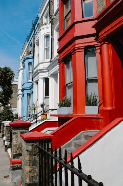 View of a row of Victorian-style terraced houses on a residential street, featuring ornate architectural details and vibrant facades in white, blue, and red. The image shows front steps made of concrete with red-painted brick walls and black wrought iron railings leading to the entrance doors. The steps are partially covered with white planter boxes containing green plants. The pavement is visible in the foreground, with a clear blue sky overhead. This setting illustrates typical urban housing applicable to home relocation or furniture transport services provided by Notting Hill Movers, highlighting the exterior environment where packing and loading processes may occur during house removals or moving projects.