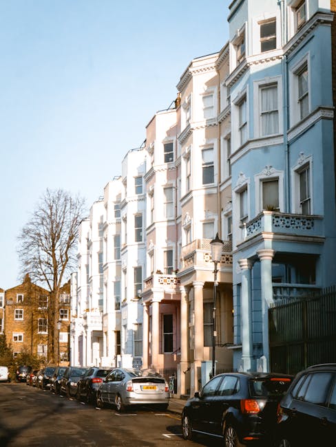 A row of multi-storey residential buildings painted in light blue and white, featuring bay windows, decorative railings, and small balconies, situated along a narrow street with parked cars lining the