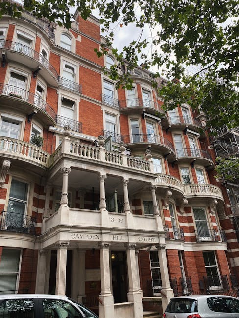 A row of multi-storey residential buildings painted in light blue and white, featuring bay windows, decorative railings, and small balconies, situated along a narrow street with parked cars lining the