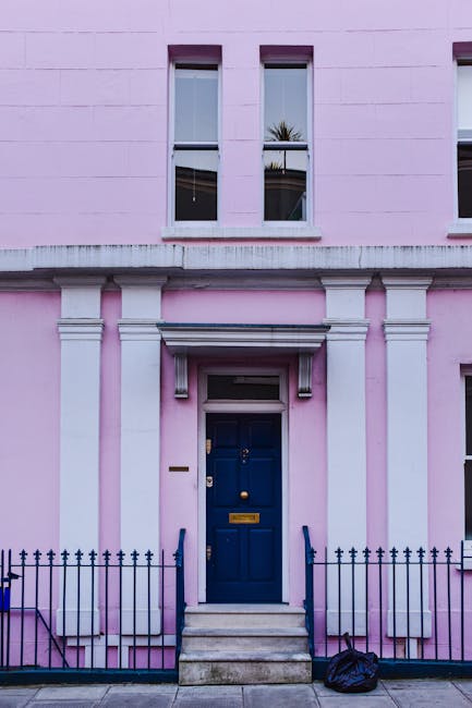 The image depicts the front entrance of a pink terraced house in W11, featuring a deep blue front door accessed by a small flight of marble steps with metal handrails on either side. The building's façade has three tall, narrow windows on the upper floor, all with white frames, and a decorative white lintel above the doorway. In front of the house is a black metal railing lining the small pavement area, and a black plastic bag is placed on the ground near the steps, indicating ongoing or completed packing or loading activities. The scene is illuminated by natural daylight, with reflections visible in the upper windows. This setting aligns with professional house removals, showcasing a typical residential entrance prepared for furniture transport during a relocation, as handled by Notting Hill Movers, experts in removals and home relocation services.