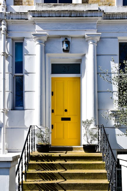 A white Victorian-style terrace house with classical architectural details, featuring a bright yellow front door, large black mailbox, and a black outdoor lantern above the door. The entrance is accessed via a short flight of stone steps with black wrought iron railings on both sides, leading from a paved pavement. Two large black planters with small shrubs flank the staircase, and a small tree is visible to the right side of the doorway. The scene is illuminated by natural daylight, emphasizing the clean, well-maintained facade. This setting reflects a typical London residential property, suitable for house removals or moving services offered by Notting Hill Movers, particularly in navigating tight stairs or narrow entrances during a home relocation.
