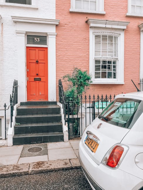 The image depicts the front entrance of a pink terraced house in W11, featuring a deep blue front door accessed by a small flight of marble steps with metal handrails on either side. The building's façade has three tall, narrow windows on the upper floor, all with white frames, and a decorative white lintel above the doorway. In front of the house is a black metal railing lining the small pavement area, and a black plastic bag is placed on the ground near the steps, indicating ongoing or completed packing or loading activities. The scene is illuminated by natural daylight, with reflections visible in the upper windows. This setting aligns with professional house removals, showcasing a typical residential entrance prepared for furniture transport during a relocation, as handled by Notting Hill Movers, experts in removals and home relocation services.