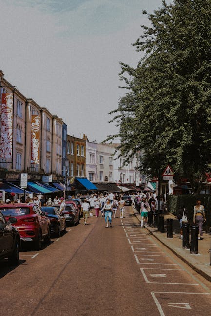 A busy street scene in Notting Hill with a line of parked cars on the left side and a large group of pedestrians walking along the pavement and crossing the road. The buildings lining the street are multi-storey, featuring colourful facades with shops and cafes on the ground level, some with striped awnings. On the right side, there are trees providing shade, and a set of bollards separating the pedestrian walkway from the roadway. The image captures a typical day of activity, with people engaged in shopping, socialising, or heading to and from local stores. This setting is relevant to house removals and moving services, illustrating an urban environment where furniture transport and relocation logistics often occur, emphasizing the importance of careful planning during packing and loading processes, especially in congested areas like Westbourne Grove. Notting Hill Movers operates in such scenarios, assisting customers with smooth and efficient home relocation efforts.