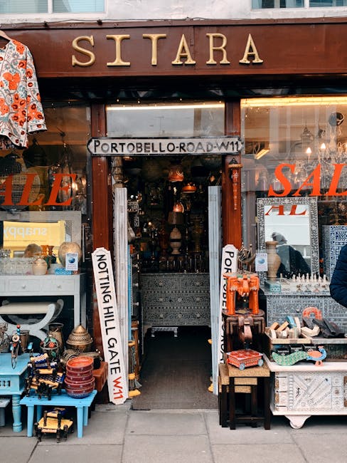 The image shows the storefront of the shop named SITARA on Westbourne Grove in Notting Hill during daylight hours. The shop's entrance is open, revealing a dimly lit interior filled with various decorative items, furniture, and lighting fixtures. Outside the shop, several tables and displays feature an assortment of objects, including small antique-style lamps, pottery, and woven accessories. To the left, a bright blue table displays miscellaneous items, while to the right, a wooden table holds stacked boxes and assorted smaller objects. Vertical signs on either side of the doorway, with one reading 'NOTTING HILL GATE W11,' indicate the location. There is a large sign with the word 'SALE' partially visible in red on the right side of the shop window, emphasizing ongoing sales. The overall scene suggests a vintage or second-hand shop that may be involved in home relocation or furniture transport services, related to moving or packing tasks supported by Notting Hill Movers, although no moving activity is directly visible in the image.